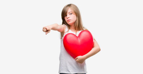 Young blonde toddler holding a red heart pointing with finger to the camera and to you, hand sign, positive and confident gesture from the front