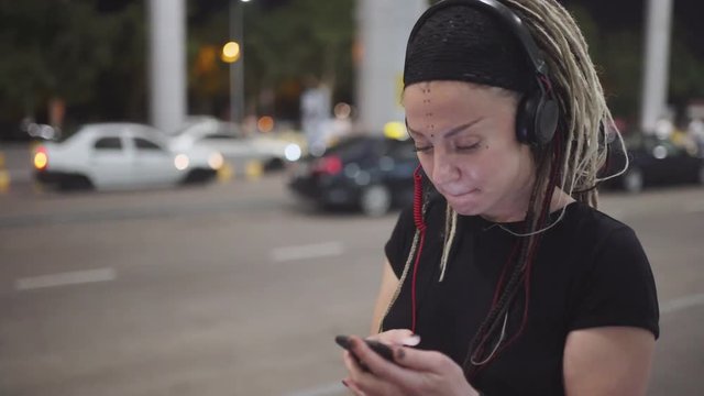 Young attractive woman with dreadlocks use smartphone listening music on headphones. On street at night 