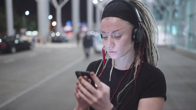 Young attractive hipster woman use smartphone listening music and dance on street at night 
