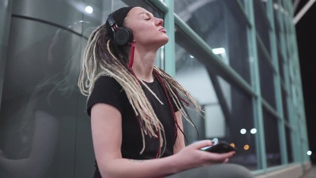 Young attractive woman with dreadlocks use smartphone listening music on headphones. On street at night 