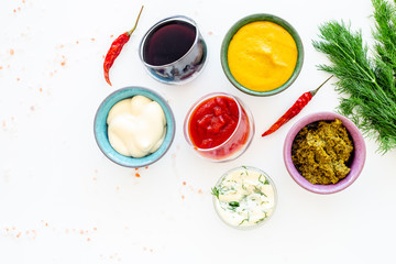 Different dip sauce in bowls near chili pepper and greenery. Ketchup, mayonnaise, mustard, soy sauce, barbecue sauce, pesto, mustard, sour on white background top view copy space
