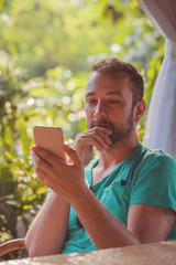 Naklejka premium Modern man sitting on wooden chair in tropical garden and using cellphone.