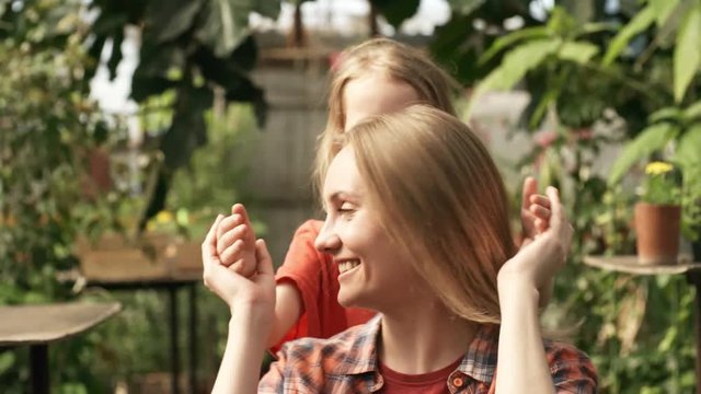 Tilt down of cute little girl surprising mother gardening in greenhouse: she is covering her eyes and hugging her from behind