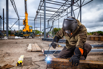 A young  man welder in brown uniform, welding mask and welders leathers, weld  metal  with a arc welding machine at the construction site, blue sparks fly to the sides - obrazy, fototapety, plakaty