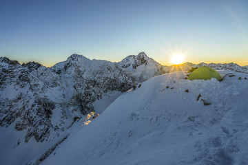 Yellow tent pitched up on a summit of a high alpine peak. Winter camping in snow covered mountains. Winter alpine landscape, climbing bivouac panorama. Vibrant blue sky, climber next to the tent.