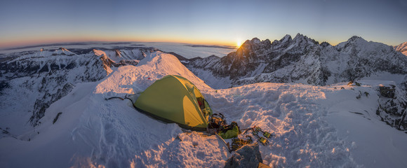 Yellow tent pitched up on a summit of a high alpine peak. Winter camping in snow covered alpine like mountains. Winter alpine landscape, climbing bivouac panorama. Vibrant blue sky, sunset or sunrise. - obrazy, fototapety, plakaty