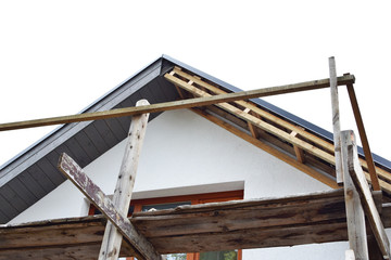 Roof construction with unfinished grey eaves and old wooden scaffolding next to renovated white  house on clear sky background with copy space. Process of installing eaves of roof and DIY concept.
