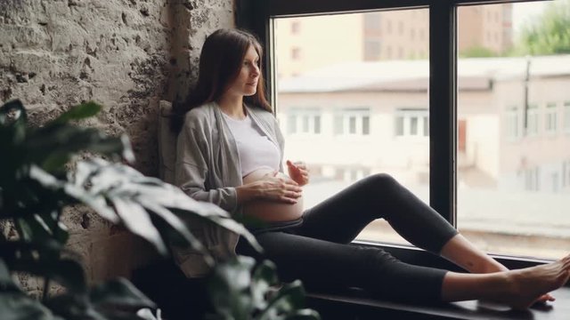 Pregnant girl beautiful brunette is sitting on window sill, stroking her belly and looking out of the window resting. Modern interiors, pregnancy and people concept.