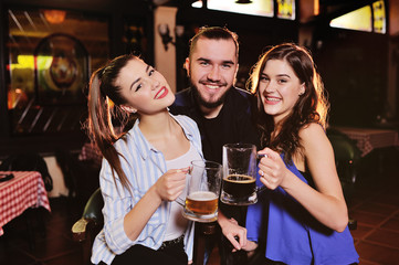 friends - young guy or man and two cute girls smiling holding glasses with beer on the background of the bar