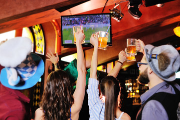 group or company of young people friends in Bavarian caps - guys and girls watching football on TV in a sports bar holding glasses with beer. Celebration of the Oktoberfest festival