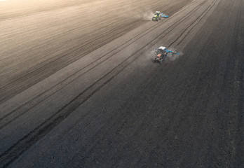 Aerial view of two blue tractors plows the earth in field on a summer day against a black earth background. Agriculture. Two tractors travel one after another along the black field