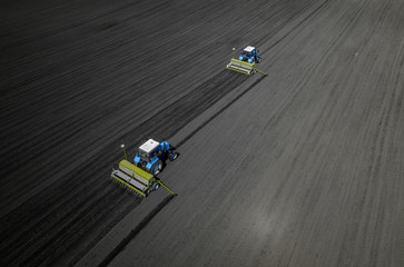 Aerial view of two blue tractors plows the earth in field on a summer day against a black earth...