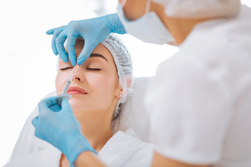 Perfect skin. Skilled experienced beautician looking at her clients face while doing an injection in the nose
