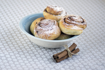 Cinnamon snails called kanelbullar, very tasty sweet rolls in blue bowl on floral pattern tablecloth, raw cinnamon