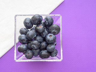 Healthy superfood blueberries in square glass bowl. Purple surface with white napkin for modern, colour block style.