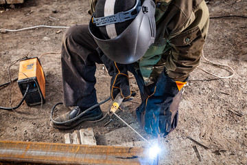 Close up man welder in welding mask, building uniform and blue protective gloves brews metal...