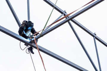 A man  welder working at height with insurance  in  welding mask is weld metal and is sitting on a metal structure at an altitude against the blue sky - Powered by Adobe