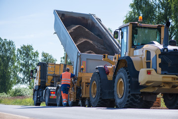 Road construction workers repairing highway road on sunny summer day. Loaders and trucks, heavy vibration roller compactor with arrow road sign on newly made asphalt.  © lainen