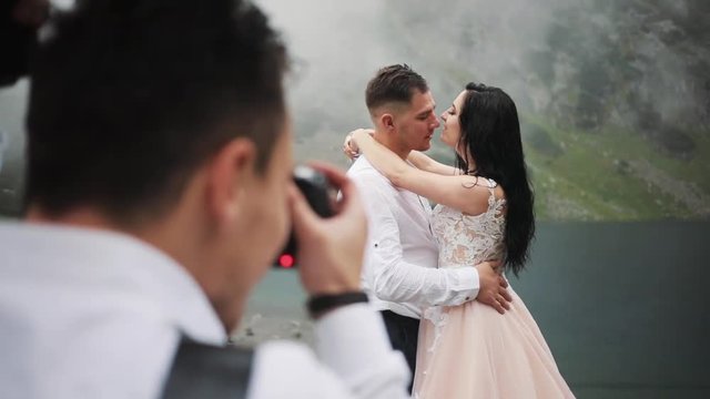 Photographer taking pictures lovely wedding couple stands near lake in mountains, hugging kissing on a cloudy day groom happy portrait dress celebration party face bridge summer young nature outdoor