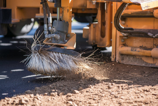 Spinning brush cleaning newly laid asphalt. Road construction workers repairing highway road on sunny summer day. Heavy machinery working on street. Road curbs being constructed with gravel