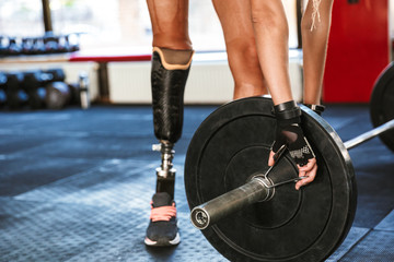 Cropped image of gymnastic young handicapped woman wearing prosthesis in tracksuit, training and lifting barbell in gym