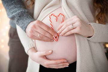 The hands of a pregnant woman and her husband hold a heart from Christmas candies on a belly background. Close up