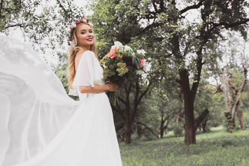 happy blonde bride in wedding dress and floral wreath holding bouquet and smiling at camera in park