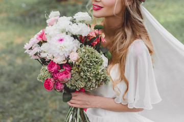 cropped shot of smiling young bride in wedding dress and veil holding bouquet of flowers outdoors