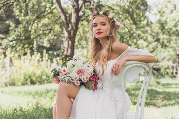 beautiful young blonde bride with wedding dress sitting on chair and looking at camera outdoors