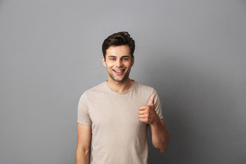 Image of attractive man 30s having stubble showing thumb up with happy smile, isolated over gray...