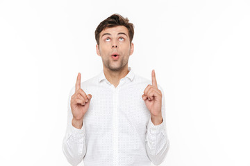 Photo of cheerful man 30s smiling and pointing fingers upward at copyspace, isolated over white...