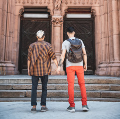 Fototapeta na wymiar Full length two males keeping each other hands while looking at beautiful doors with statue of old building. They turning back to camera