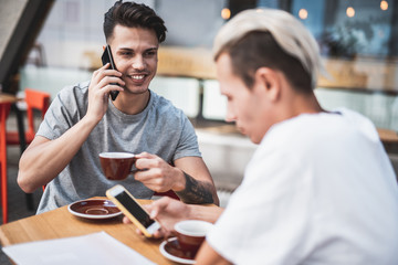Smiling man talking on phone while spending time with pensive friend. He typing in gadget at table in cafe