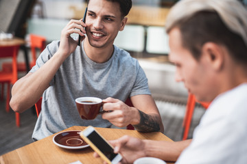 Positive male speaking on phone while tasting cup of liquid. He locating opposite thoughtful comrade using digital device