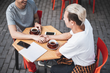 Top view outgoing man holding hand of positive friend while sitting at desk. They tasting mugs of beverage and using phones during conversation