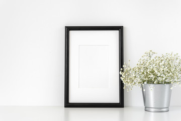 Black a4 frame mockup in interior. Frame mock-Up poster or photo frame and supplies and vase with flowers on table near white wall. Desk space, copy space. Background