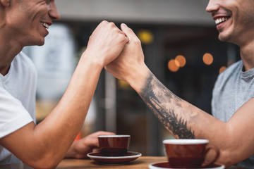 Beaming man holding hand of another guy while talking with him. Smiling comrades drinking mugs of delicious coffee while situating at table in cafe