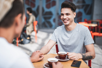 Portrait of laughing guy telling with comrade while looking at him and holding his hand. They having leisure at table while drinking mugs of beverage