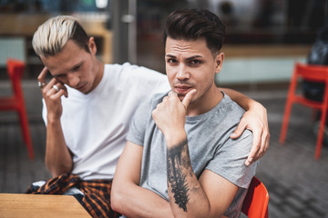 Thoughtful man looking at camera while keeping head during communication with contemplative comrade. He hugging him while sitting at table in cafe outside