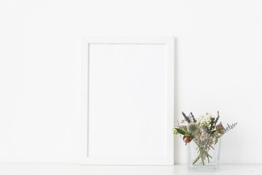 Minimal white a4 portrait frame mockup with dried field wild flowers in transparent vase on white wall background. Empty frame, poster mock up for presentation design. 