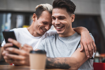 Obraz premium Portrait of laughing guy hugging outgoing man while watching at phone. He typing in appliance during communication with comrade