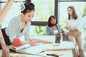 Obraz premium Smiling young asian businesswoman working with blueprint while colleagues sitting behind