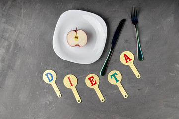 The word "diet" is collected from a children's educational card with letters. Unusual black cutlery, fork and knife ready for breakfast, lunch, dinner. Half red apple on a plate, too little food.