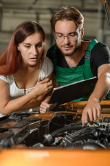 A young mechanic shows the cause of the malfunction of an orange car of the girl client