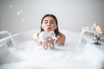 Portrait of serene woman blowing foam from hands while locating in warm water during rest in bath