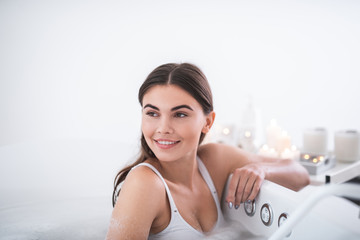 Portrait of glad girl with attractive smile leaning on side of bath while having spa treatment procedure with foam water