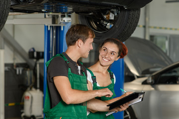 A beautiful and young girl locksmith conducts diagnostics of the suspension of the car with her colleague from the car-care center, they are dressed in green overalls
