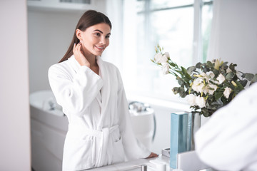 Portrait of satisfied beautiful female watching at reflection while locating near sink in bathroom