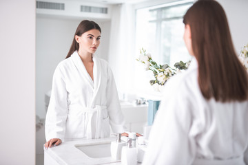Fototapeta premium Portrait of calm pretty girl looking at mirror. She wearing cozy bathrobe while locating in apartment