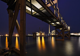 Bremen, Germany - Brightly lit harbor at night with rusty steel jetty above, yellow lights reflecting in the water and various industrial plants in the distance